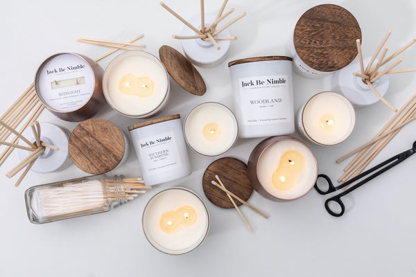 Flat lay of Jack Be Nimble soy candles and reed diffusers with wooden lids, featuring scents like Woodland and Midnight, displayed with wick trimmers and matchsticks.