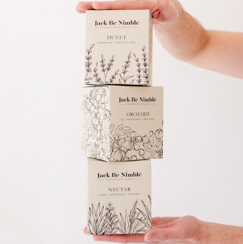 Three 'Jack Be Nimble' candle boxes held by a hand on a white background