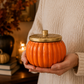 A person in a cream knit sweater holds a ribbed orange ceramic jar with a gold lid in a warm, cozy indoor setting.