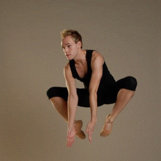 A male dancer in a black sleeveless top and black fitted pants leaps mid-air against a plain warm beige studio background.